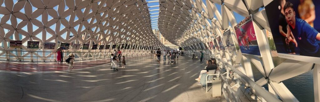 Astana bridge view: People walking through a modern, covered bridge with geometric patterns and photo displays.